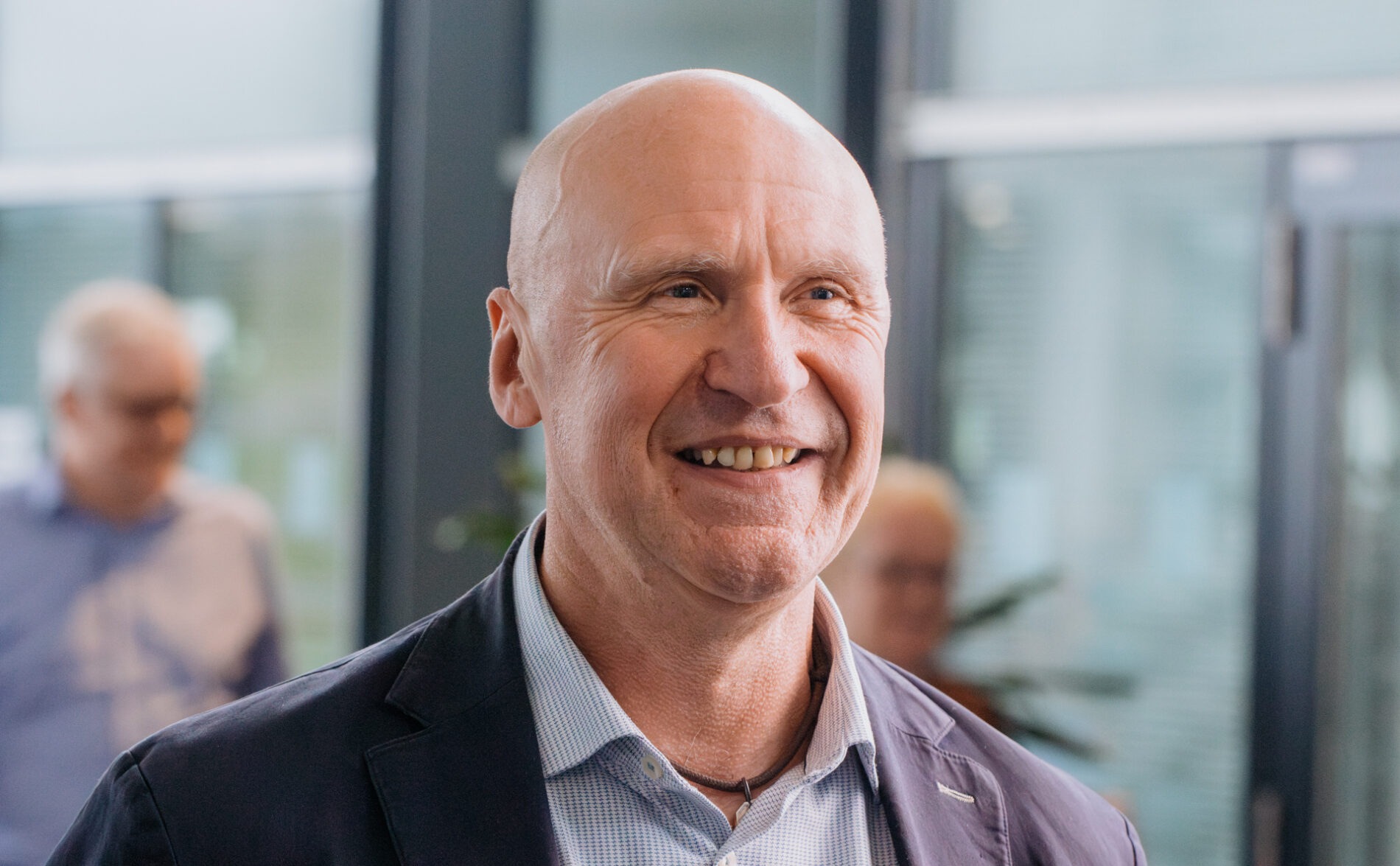 A head-and-shoulders portrait of a smiling bald man in a blue suit jacket and checked white and blue shirt.
