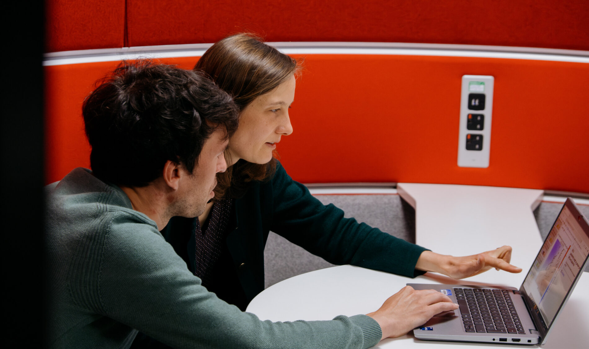 A woman with shoulder length light brown hair and a man with dark brown hair sitting at a table together looking at a laptop.