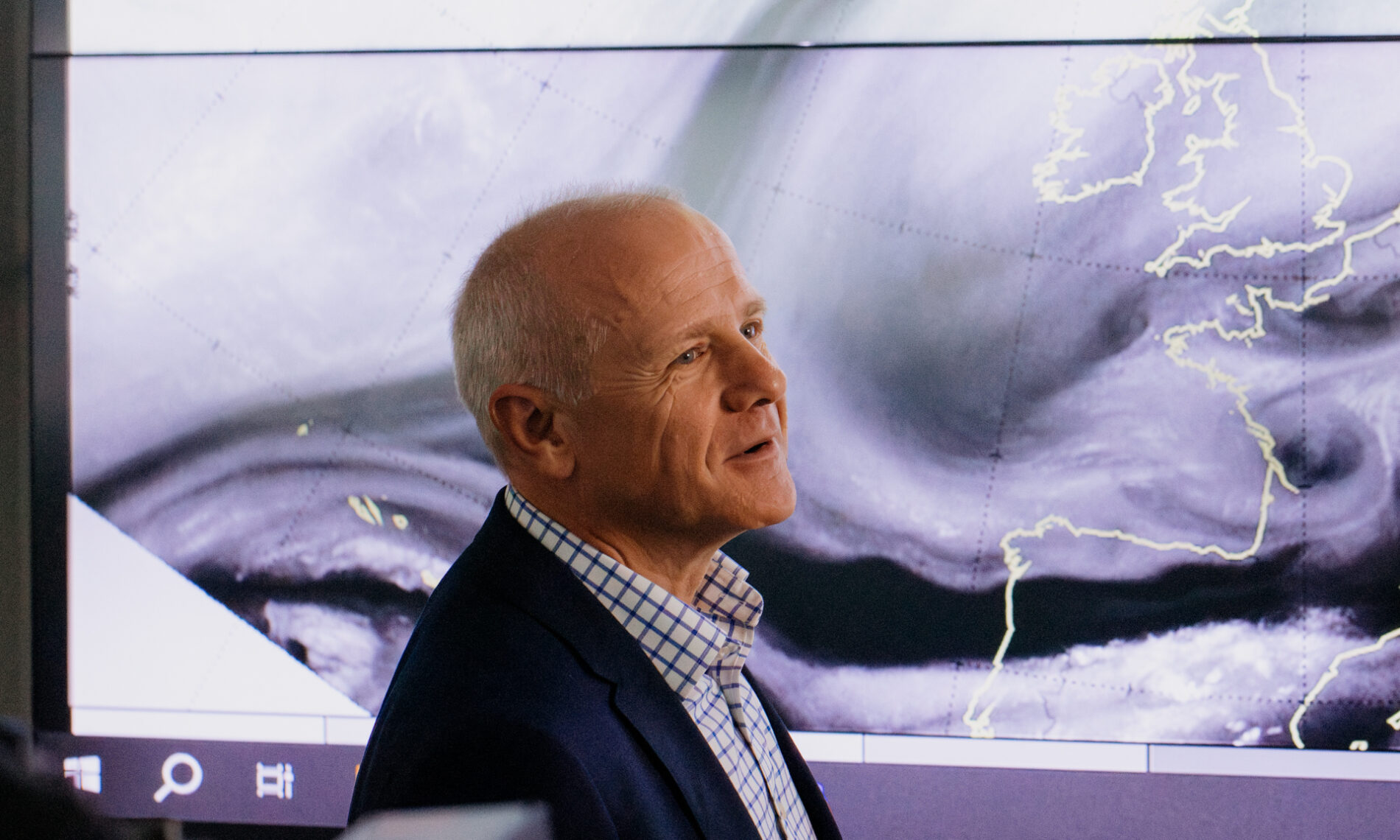 A bald man with a serious expression looking at a giant screen showing cloud formations over England and western Europe.